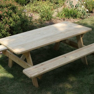 May include: A light-colored wooden picnic table with attached benches. The table has a rectangular top and is supported by angled legs. The benches are also made of wood and are attached to the table. The table is outdoors on a grassy lawn.