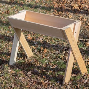 May include: A wooden planter box with a rectangular trough and angled legs. The natural wood tones create a rustic aesthetic. The planter is designed for outdoor use and is set on a bed of leaves and grass.