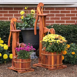 May include: Two wooden water pump planters with flowers. The planters are made of stained wood and have a bucket-like design. One planter holds purple flowers, and the other holds yellow flowers. The background includes a brick wall and green bushes.