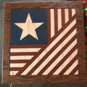 May include: A wooden framed wall art piece featuring a stylized American flag design with red, white, and blue colors. The flag is made of wood and has a raised star in the upper left corner.
