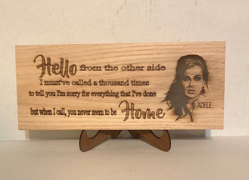 Może przedstawiać: Drewniany znak z laserowo grawerowanym obrazem Adele i tekstem z jej piosenki "Hello". Tekst brzmi: "Hello from the other side I must've called a thousand times to tell you I'm sorry for everything that I've done but when I call, you never seem to be Home".