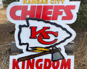 May include: White wooden sign with the words "Kansas City Chiefs Kingdom" in red letters. The sign also features a red and black "KC" logo inside a white arrowhead shape.