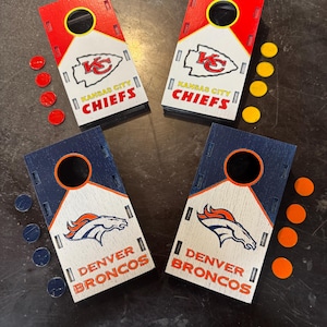 May include: Four miniature cornhole game boards, two featuring the Kansas City Chiefs logo and two with the Denver Broncos logo. Each board has a circular hole and is accompanied by colored bean bags. The boards are red, white, and blue.