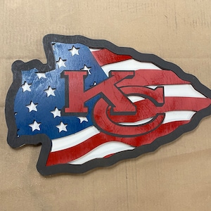 May include: A wooden arrowhead-shaped sign with a red, white, and blue American flag design. The letters "KC" are cut out of the flag and painted red. The sign has a black border.