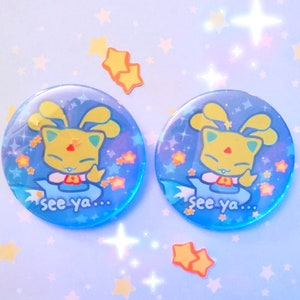 May include: Two blue and yellow round buttons with a cartoon character on each. The character is a yellow cat with blue eyes and a pink nose. The character is wearing a blue and white outfit and has a blue halo. The buttons have a blue background with white stars and the text "see ya..." on each.