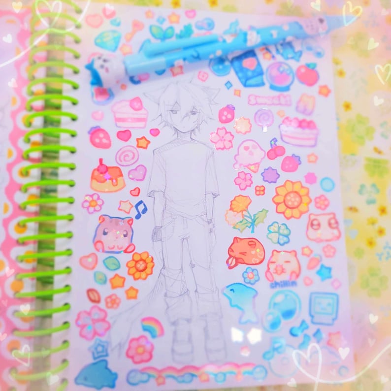 May include: A white notebook with a spiral binding and a pencil on top. The notebook is covered in colorful stickers, including hearts, stars, flowers, and cartoon characters. There is a pencil drawing of a person on the page.