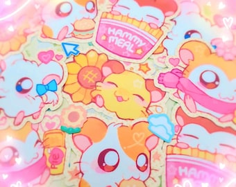 Op de afbeelding: Een verzameling kleurrijke stickers met cartoonhamsters. De stickers tonen hamsters met verschillende accessoires, waaronder hamburgers, friet en strikken. Sommige stickers hebben de tekst "Hammy Meal". De stickers zijn in tinten oranje, roze, geel en blauw.