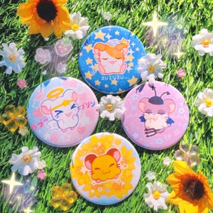 May include: Four round button pins featuring different cartoon hamsters. The first pin is blue with white stars and a hamster wearing a crown and halo. The second pin is pink with a haloed hamster with a crown and wings. The third pin is pink with a hamster wearing a black hat and devil horns. The fourth pin is yellow with a hamster wearing a crown and halo.