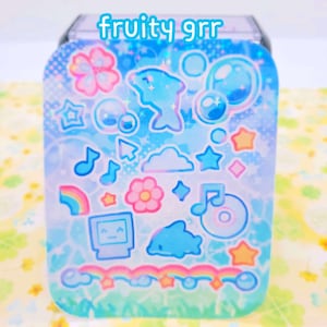 May include: A blue and green holographic sticker sheet with a variety of cute stickers, including dolphins, stars, rainbows, clouds, and music notes. The text "fruity grr" is printed on the sheet.