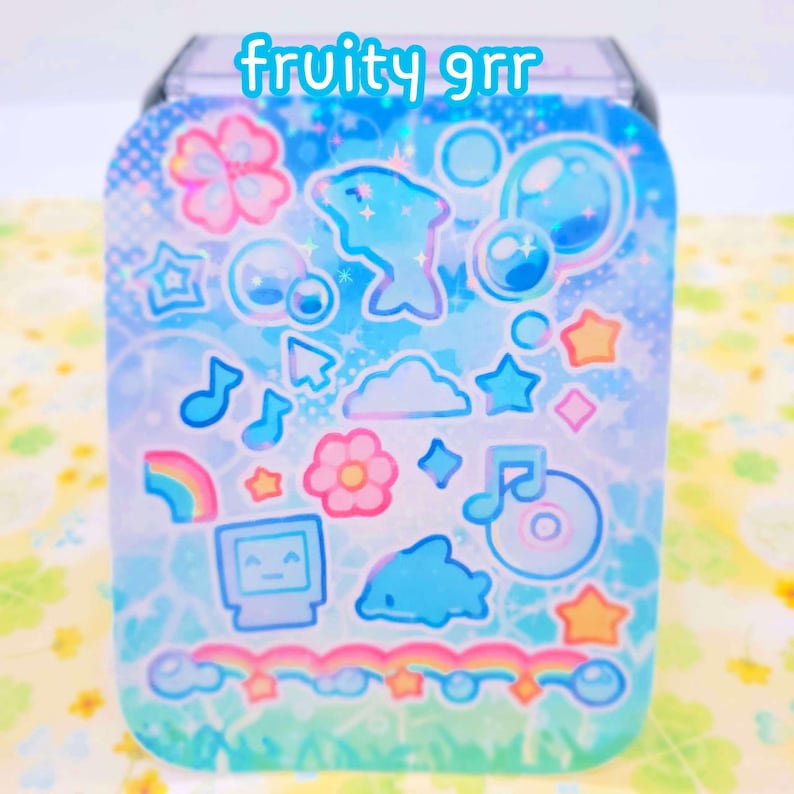 May include: A blue and green holographic sticker sheet with a variety of cute stickers, including dolphins, stars, rainbows, clouds, and music notes. The text "fruity grr" is printed on the sheet.