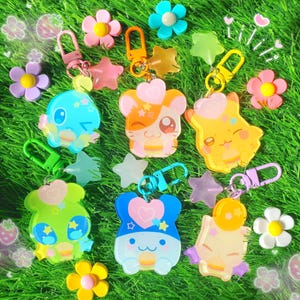 May include: A collection of colorful acrylic keychains featuring various cartoon characters, stars, and flower designs. The keychains have gold, green, and purple clasps. The background is green grass.