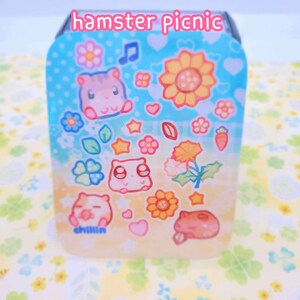 May include: A blue and yellow square container with a whimsical design featuring cartoon hamsters, flowers, stars, and the text "hamster picnic" and "chillin".