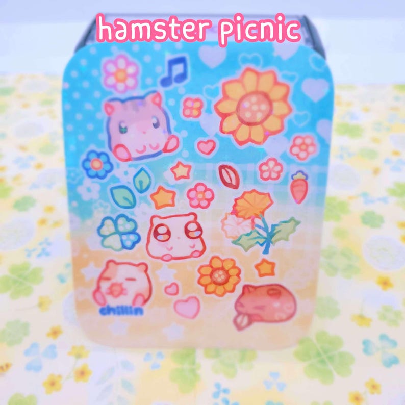 May include: A blue and yellow square container with a whimsical design featuring cartoon hamsters, flowers, stars, and the text "hamster picnic" and "chillin".