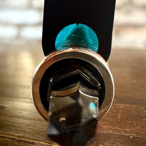 K&ouml;nnte beinhalten: Ein silberner Ring mit einem klaren Kristallstein in der Mitte. Der Kristall ist pyramidenf&ouml;rmig mit einer sechseckigen Basis. Der Ring wird von einem Finger gehalten, und eine schwarze Karte mit einem t&uuml;rkisfarbenen Kreis befindet sich hinter dem Ring.