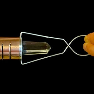 K&ouml;nnte beinhalten: Ein kupferfarbener Metallstab mit einem klaren Kristall, der in einer Drahtschlaufe gehalten wird. Die Drahtschlaufe wird von einer Hand gehalten.