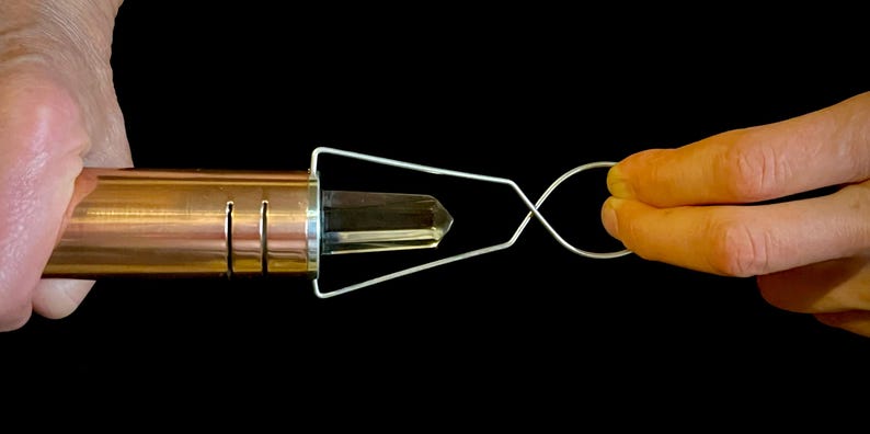 K&ouml;nnte beinhalten: Ein kupferfarbener Metallstab mit einem klaren Kristall, der in einer Drahtschlaufe gehalten wird. Die Drahtschlaufe wird von einer Hand gehalten.