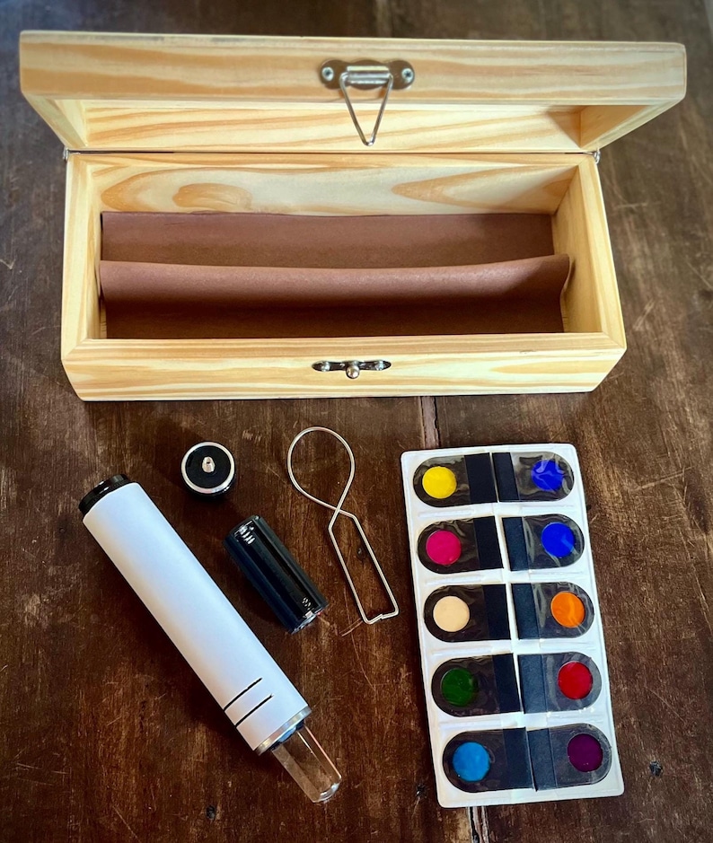 K&ouml;nnte beinhalten: Eine Holzkiste mit einem wei&szlig;en und durchsichtigen Glasrohr, einer schwarzen Batterie, einem Metallring und einer Plastikfolie mit acht farbigen Kreisen. Die Kreise sind gelb, rosa, beige, gr&uuml;n, blau, rot, orange und lila.