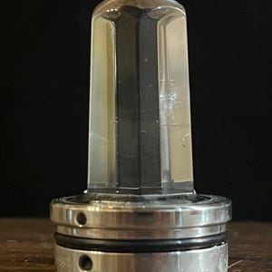 K&ouml;nnte beinhalten: Ein durchsichtiger Glasflaschenverschluss mit einem silbernen Metallsockel. Der Verschluss hat eine sechseckige Form und der Sockel hat zwei Ringe.