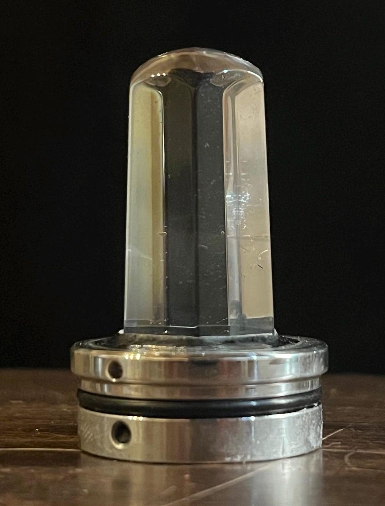 K&ouml;nnte beinhalten: Ein durchsichtiger Glasflaschenverschluss mit einem silbernen Metallsockel. Der Verschluss hat eine sechseckige Form und der Sockel hat zwei Ringe.