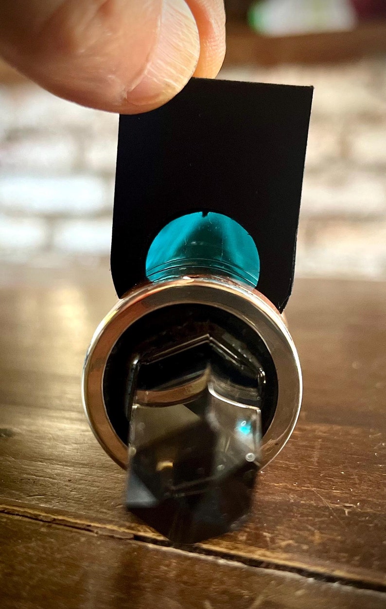 K&ouml;nnte beinhalten: Ein silberner Ring mit einem klaren Kristallstein in der Mitte. Der Kristall ist pyramidenf&ouml;rmig mit einer sechseckigen Basis. Der Ring wird von einem Finger gehalten, und eine schwarze Karte mit einem t&uuml;rkisfarbenen Kreis befindet sich hinter dem Ring.