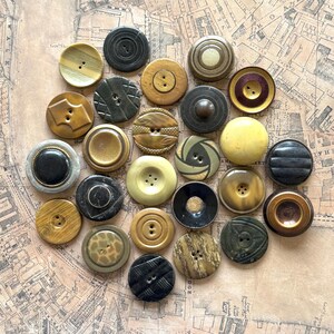 May include: A collection of twenty-four round buttons in various shades of brown, tan, black, and olive green. The buttons feature diverse textures, including smooth, grooved, patterned, and rimmed designs, with two or four holes for attachment.
