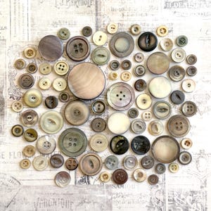 May include: A collection of vintage buttons in various sizes and shades of cream, brown, and gray. The buttons are made of different materials, including mother-of-pearl, and feature two or four holes. The buttons are arranged on a patterned background.