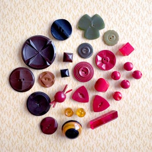 May include: A collection of assorted buttons in various shapes, sizes, and colors including dark brown, black, olive green, red, and amber. The assortment features round, square, triangular, and rectangular buttons with two or four holes.