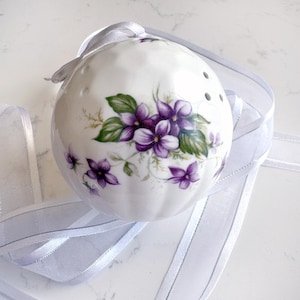 May include: A spherical white ceramic ornament featuring a painted floral design of purple violets with green leaves. The surface has a dimpled texture and small holes near the top. A sheer white ribbon is threaded through the top for hanging.