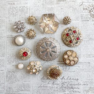 May include: A collection of vintage buttons in various shapes, sizes, and designs. The buttons include clear faceted glass, pearl, and ornate metalwork with rhinestones and red accents. The background is a vintage newspaper.