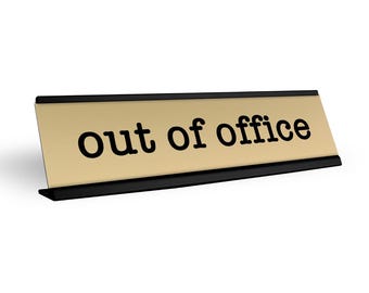 May include: A gold desk sign with black trim and the text "out of office" in black.