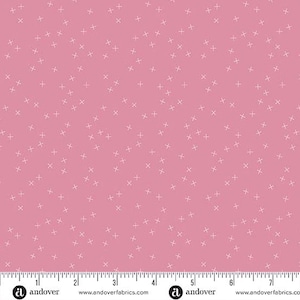 May include: Pink fabric with a pattern of small white crosses scattered across the surface. The fabric has a ruler along the bottom edge, marked in inches. The fabric is from Andover Fabrics.