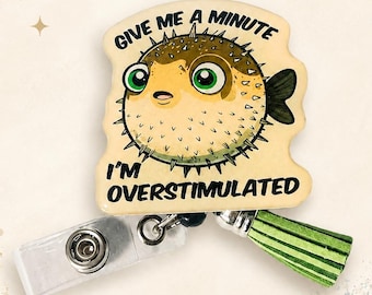 May include: A badge reel featuring a tan, round pufferfish illustration with green eyes and spikes. The text reads: GIVE ME A MINUTE I'M OVERSTIMULATED. It includes a clear plastic clip and a small, light green fabric tassel attached to the base.