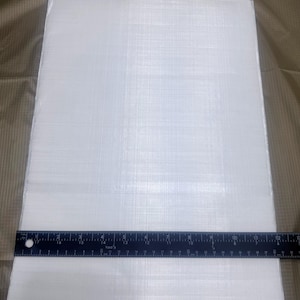 May include: A rectangular, white, textured sheet with a ruler placed across the bottom. The ruler is black with white markings and measures in inches. The sheet appears to be made of a woven material.