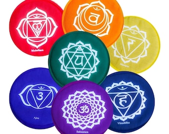Op de afbeelding: Zeven ronde, gekleurde viltkussens met witte symbolen en Sanskriet-tekst. De kleuren zijn rood, oranje, geel, groen, blauw, paars en wit. De tekst bevat "Muladhara", "Svadhisthana", "Manipura", "Anahata", "Ajna", "Vishuddha" en "Sahasrara".