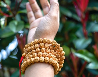 May include: A person wearing a brown beaded bracelet with a red string tied around it. The bracelet is made of small, round beads and is worn on the wrist. The person's hand is raised in the air, with the fingers spread out.