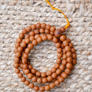May include: A brown wooden mala bead necklace with a yellow string tassel. The beads are round and have a natural wood finish. The necklace is lying on a woven rug.