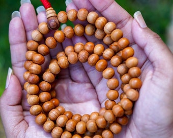 May include: A wooden mala bead necklace with a red tassel, held in cupped hands. The beads are a light brown color and are strung together in a circular shape.