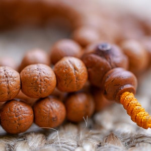 May include: A close-up of a brown wooden mala bead necklace with a yellow cord. The beads are round and have a textured surface. The necklace is lying on a woven surface.