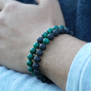 May include: Two beaded bracelets, one with green malachite beads and the other with black lava stone beads. The bracelets are worn on a wrist, showcasing the contrasting colours and textures of the natural stones. The bracelets are a stylish accessory.