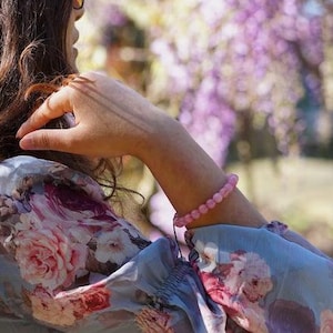 Peut inclure: Une femme portant une blouse à imprimé floral avec une sangle en cuir marron et un bracelet en perles roses. Elle se tient devant un fond de fleurs violettes.