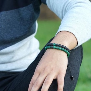 Puede incluir: Dos pulseras de cuentas en la muñeca de una persona. La pulsera superior está hecha de cuentas de lava negra y la pulsera inferior está hecha de cuentas de malaquita verde.