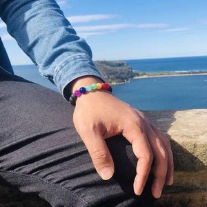 May include: A person wearing a denim shirt and black jeans with a colorful beaded bracelet on their wrist. The bracelet has black, purple, blue, green, yellow, orange, and red beads.