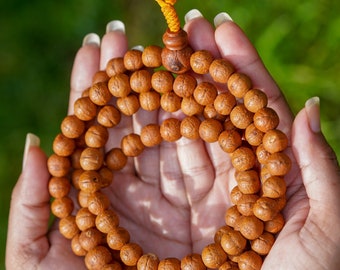 May include: A brown wooden mala bead necklace with a yellow tassel. The necklace is held in two hands.