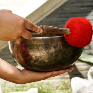 May include: A person holding a traditional Tibetan singing bowl with intricate engravings and a red felt mallet. The bowl is made of metal and has a deep, resonant sound.