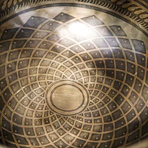 May include: A close-up of the inside of a singing bowl with a detailed geometric design. The bowl is made of metal and has a circular shape. The design is intricate and symmetrical, with a central circle surrounded by concentric rings of smaller circles.