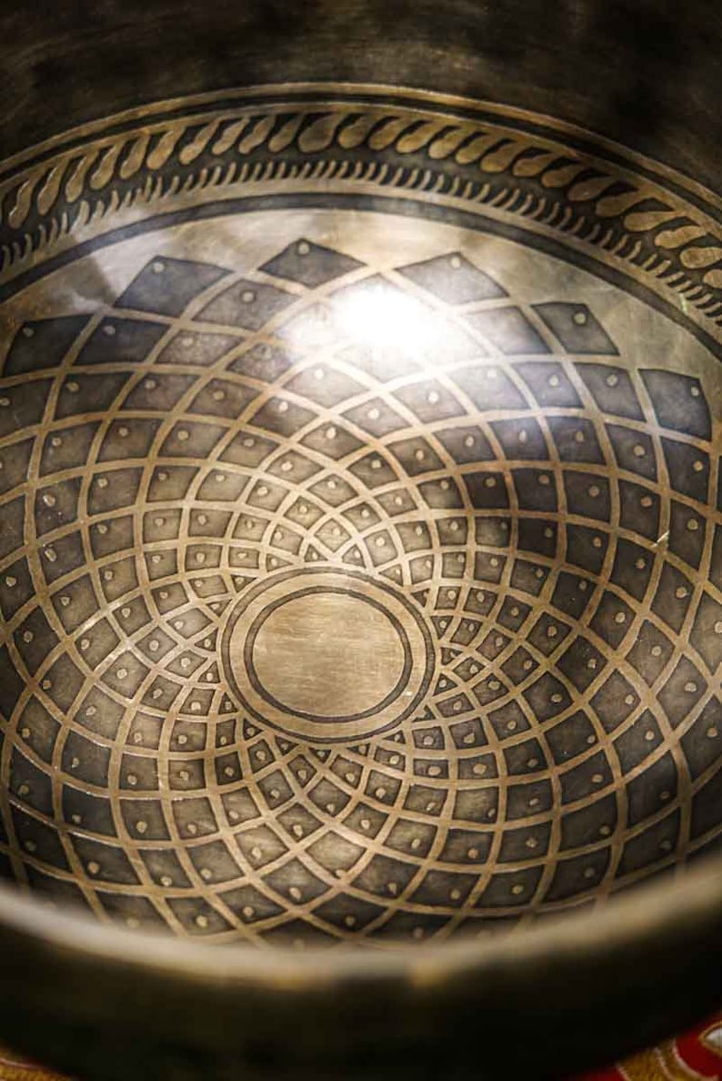May include: A close-up of the inside of a singing bowl with a detailed geometric design. The bowl is made of metal and has a circular shape. The design is intricate and symmetrical, with a central circle surrounded by concentric rings of smaller circles.