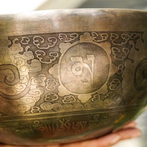 May include: A traditional Tibetan singing bowl with intricate engravings, including a central symbol and a border of swirling patterns. The bowl is made of metal and has a warm, aged patina.