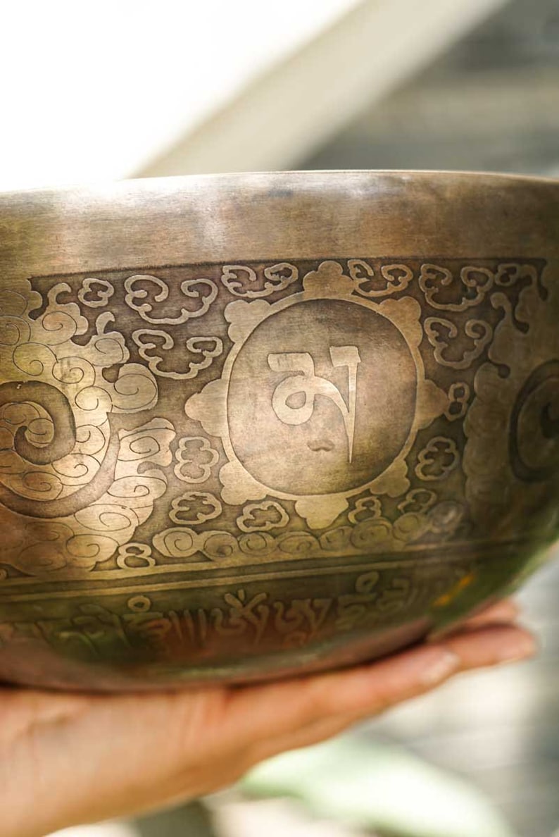 May include: A traditional Tibetan singing bowl with intricate engravings, including a central symbol and a border of swirling patterns. The bowl is made of metal and has a warm, aged patina.