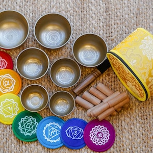 Op de afbeelding: Een set van zeven gouden zangkommen met een geel, gepatroneerd kussen en zeven chakra-kleurige ronde stickers. De kommen zijn in een cirkel gerangschikt met het kussen in het midden. De stickers zijn in een rij onder de kommen gerangschikt.