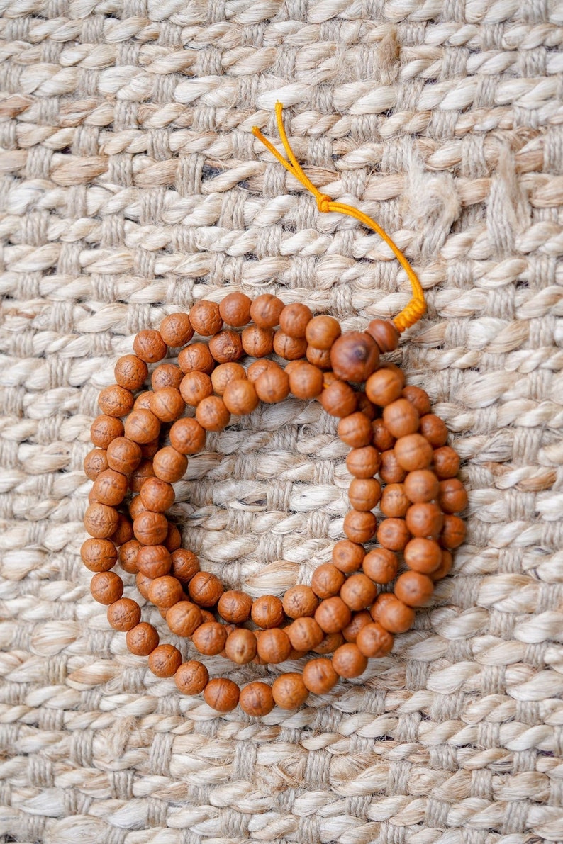 May include: A brown wooden mala bead necklace with a yellow string tassel. The beads are round and have a natural wood finish. The necklace is lying on a woven rug.